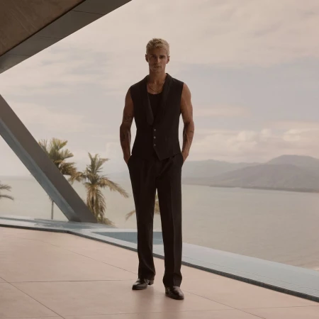Attractive adult male in a vest and black suit stands in front of tropical ocean and palm tree landscape. Fashion editorial.