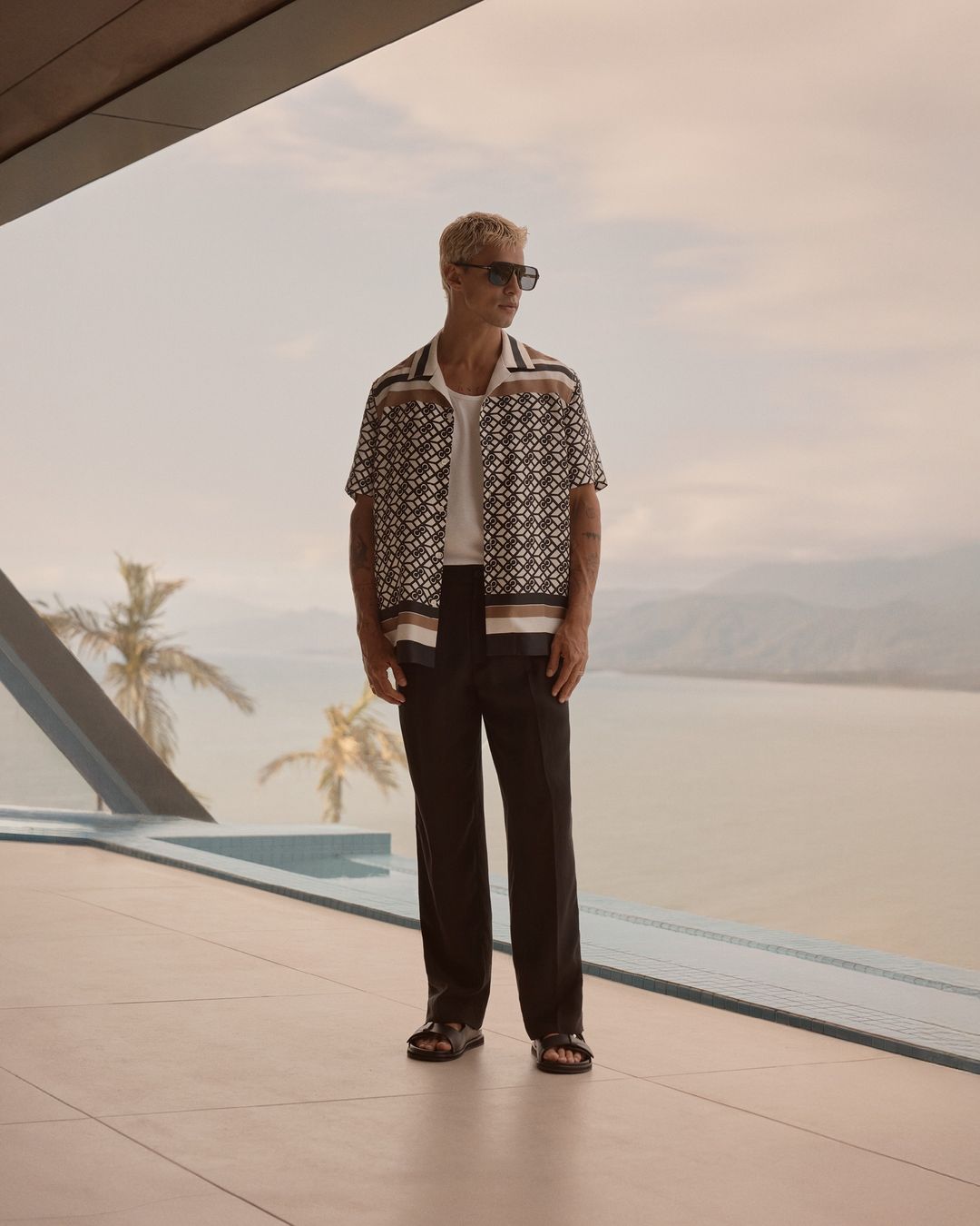 A blond male model poses in a printed shirt and black pants, set against a scenic ocean backdrop on a sunny day in a lifestyle fashion shot.