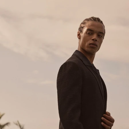 Man with braided hair wearing a dark textured blazer looking over his shoulder against an overcast sky.