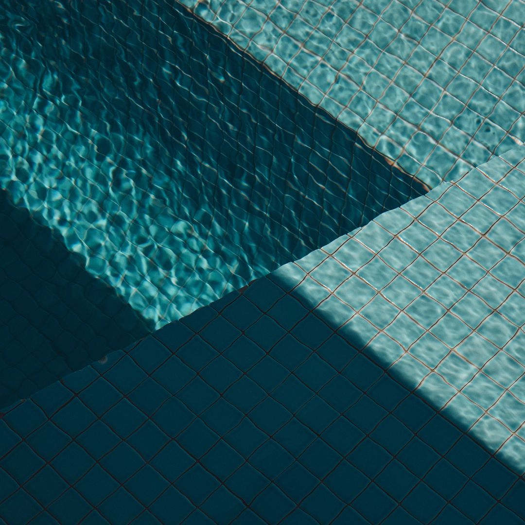 Overhead view of turquoise swimming pool water and tiled steps with geometric shadow patterns on a sunny day, creating a refreshing scene.