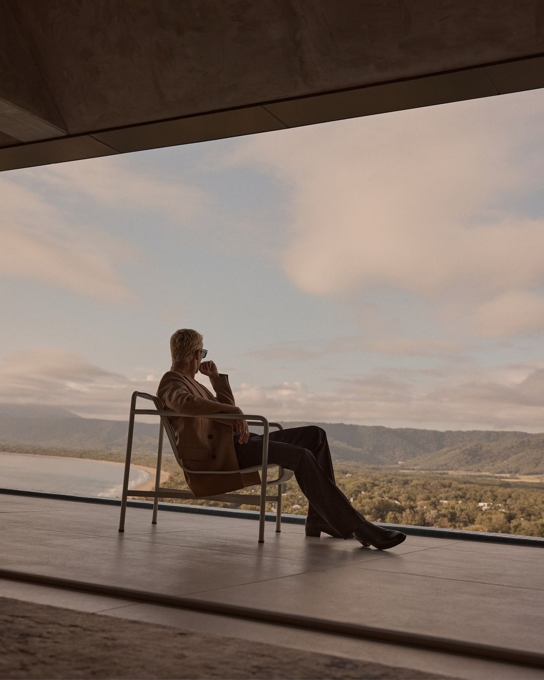 Adult in a brown suit sits on a chair overlooking trees, mountains, and cloudy sky in a tranquil outdoor setting with modern architecture.