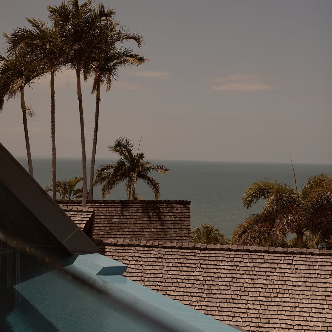 Tranquil daytime scene features an outdoor swimming pool, a shingled roof, palm trees, and a serene ocean backdrop with a clear, blue sky.