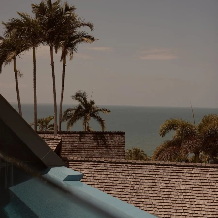 Tranquil daytime scene features an outdoor swimming pool, a shingled roof, palm trees, and a serene ocean backdrop with a clear, blue sky.
