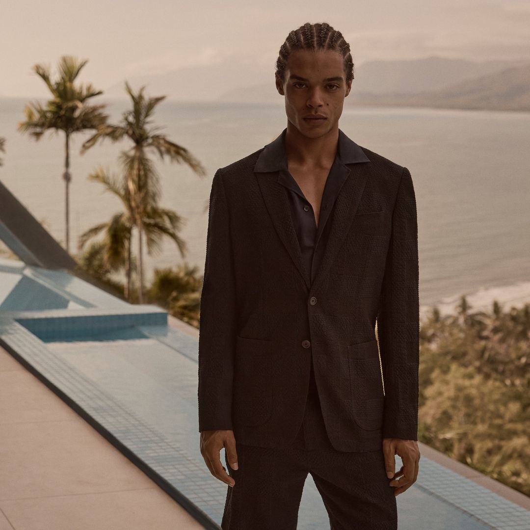 Fashion shot of a Black male model in a dark suit against a seascape backdrop, capturing elegance and style in a scenic setting.