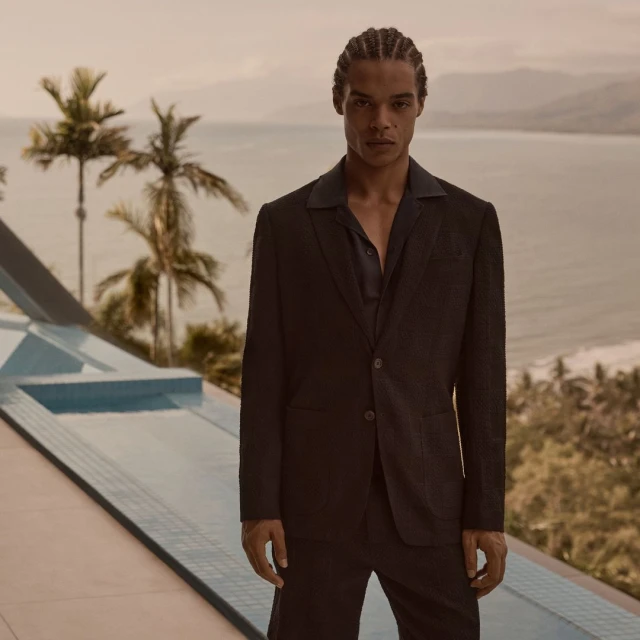 Fashion shot of a Black male model in a dark suit against a seascape backdrop, capturing elegance and style in a scenic setting.
