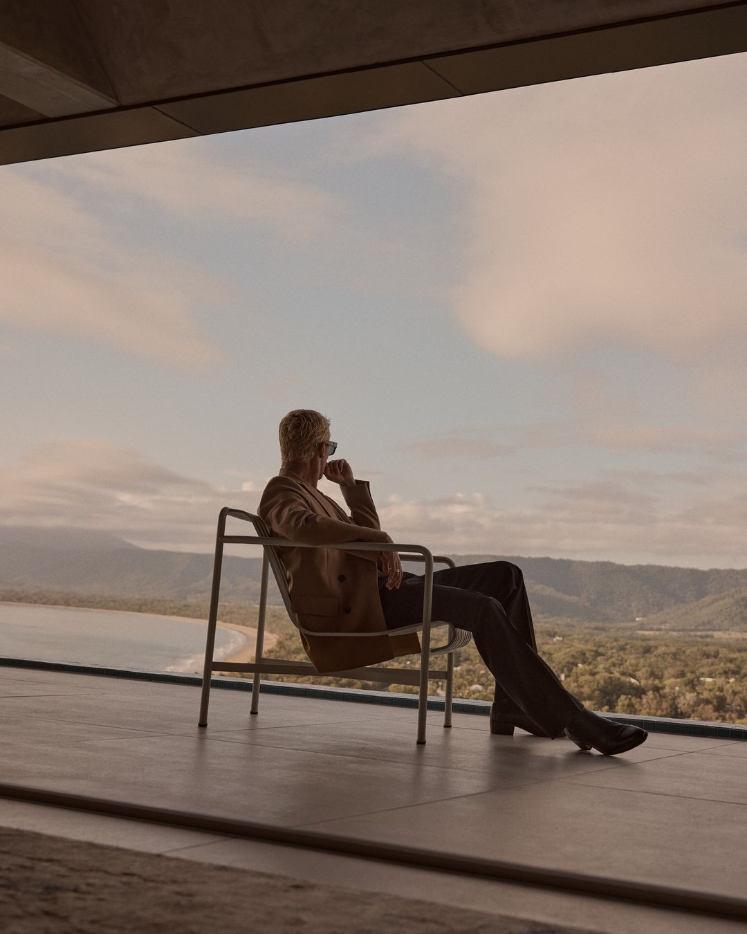 Adult relaxing in modern chair on balcony overlooking tropical coastline with forest and cloudy sky in neutral, desaturated tones.