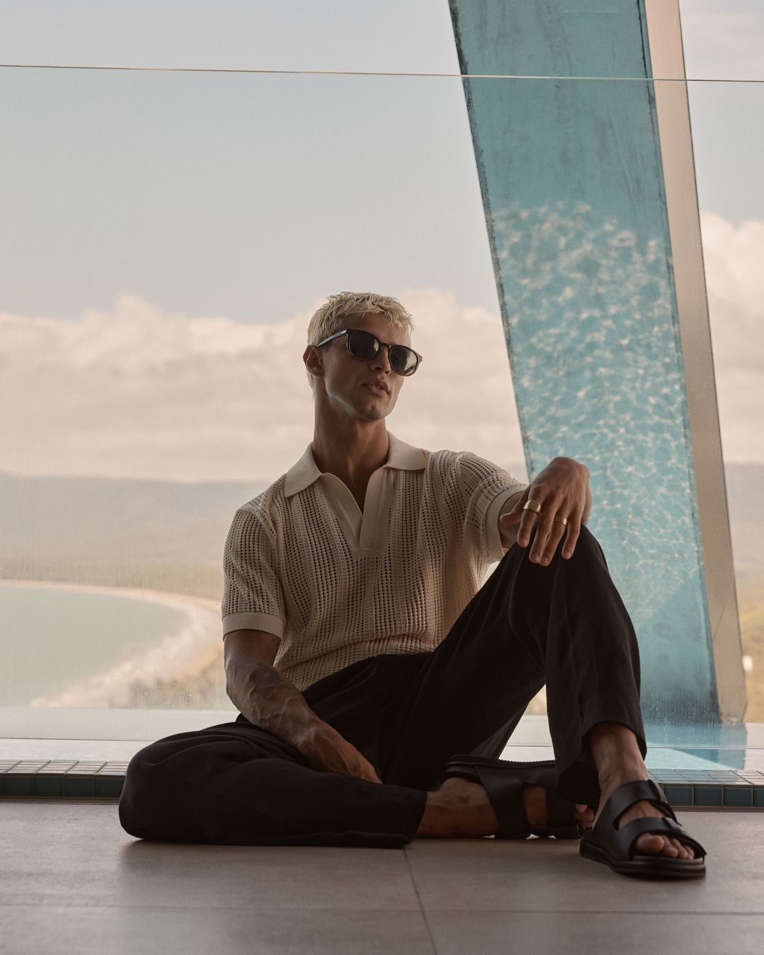 Man sitting near pool with coastal view. He is wearing sunglasses, a polo shirt, and sandals, posing relaxed in a modern setting.