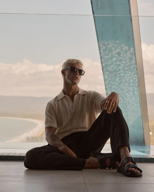 Man sitting near pool with coastal view. He is wearing sunglasses, a polo shirt, and sandals, posing relaxed in a modern setting.