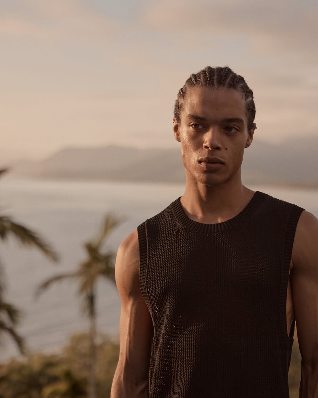 Portrait of an adult male wearing a black tank top outside with palm trees and water in the background, shot with natural light.