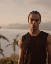 Young man with braided hair wearing a black sleeveless knit top standing outdoors against an ocean and palm tree background at sunset