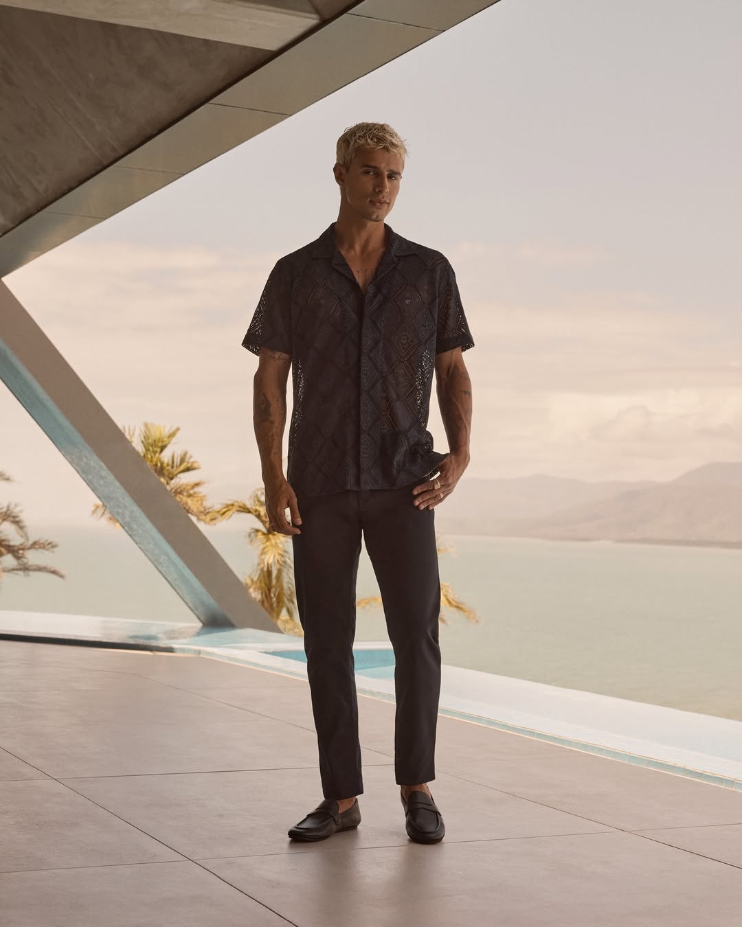 Full body shot of a blond male model in a black patterned shirt and black pants posing by a modern building overlooking a sea view.
