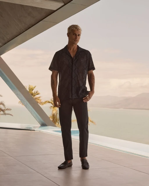 Full body shot of a blond male model in a black patterned shirt and black pants posing by a modern building overlooking a sea view.