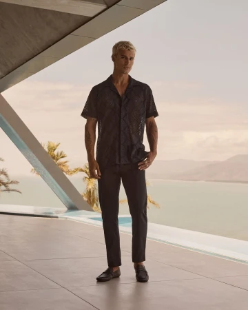 Full body shot of a blond male model in a black patterned shirt and black pants posing by a modern building overlooking a sea view.