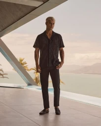 Blonde man wearing a dark patterned short-sleeve shirt and trousers standing on a modern outdoor terrace overlooking water.