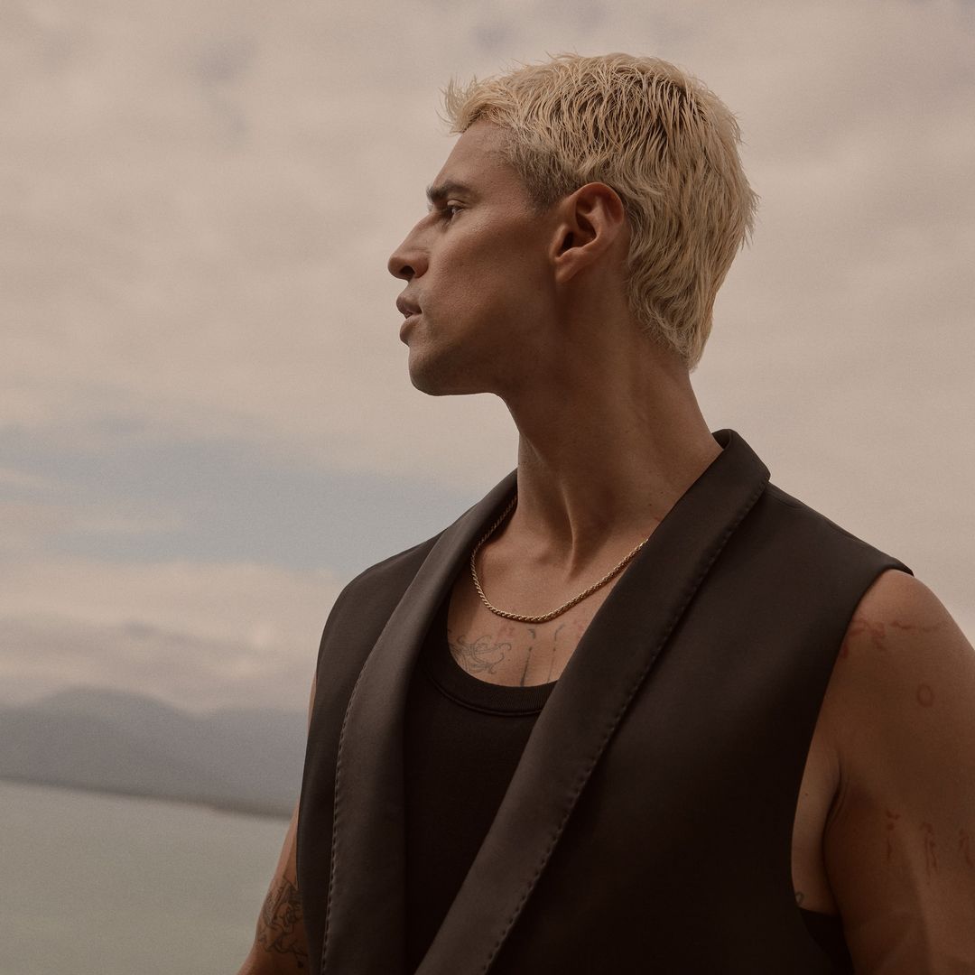 Portrait of a fair-skinned man with platinum blonde hair and a black vest standing outdoors with a water and horizon backdrop.