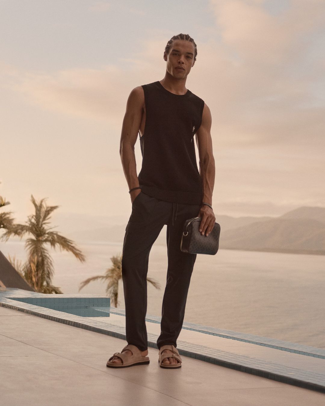 A handsome dark-skinned man poses on a patio in a black tank top and dark pants with a scenic background and a neutral color palette.