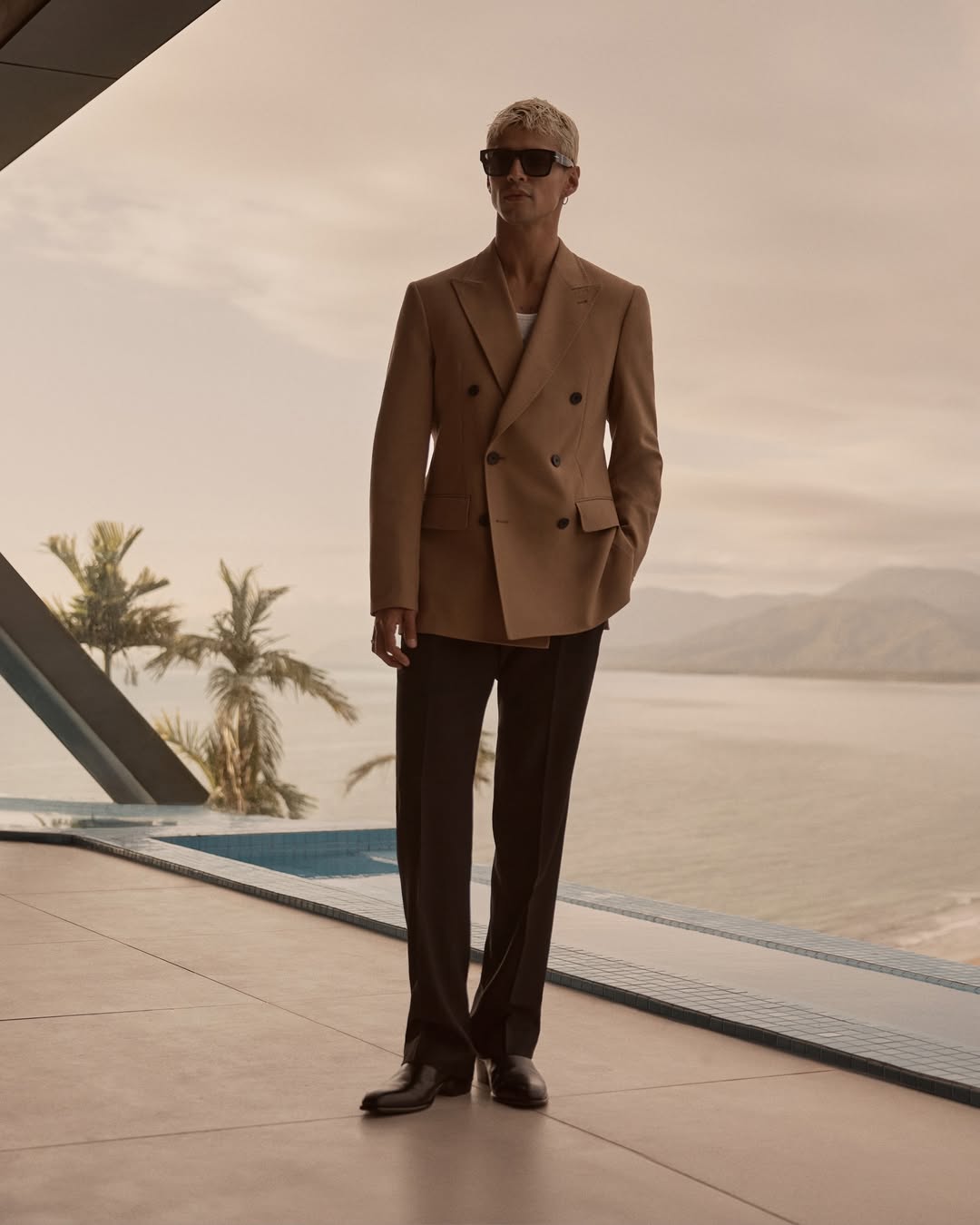 Man in a camel double-breasted blazer and dark sunglasses standing on a balcony overlooking water and palm trees