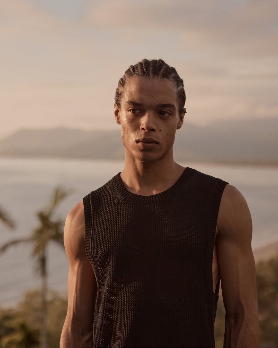 Portrait of a muscular adult male in a black sleeveless top, outdoors with a blurred coastal landscape and palm trees in the background.