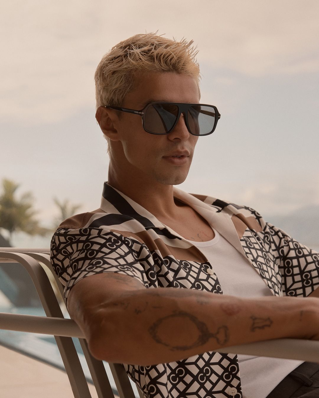 Adult male sitting outdoors wearing sunglasses and patterned shirt, looking cool and relaxed on a bright day with mountain backdrop.