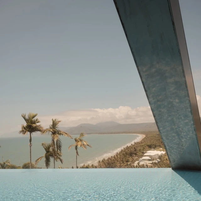 Coastal view from infinity pool featuring palm trees, sandy beach, ocean, mountains, and blue sky in tropical setting.