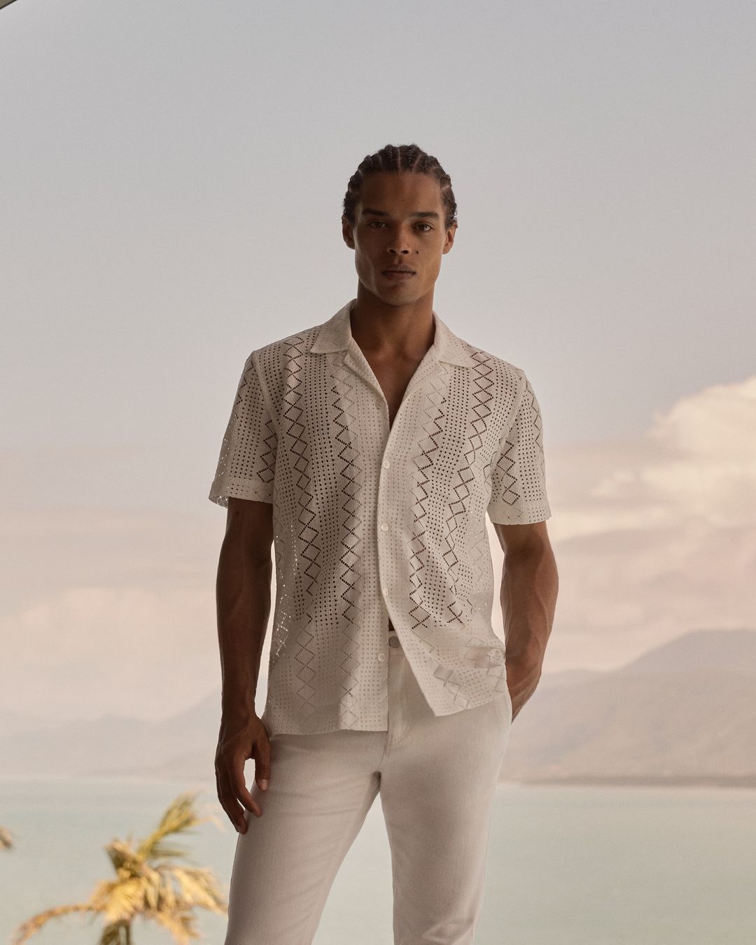 Man in white patterned shirt and pants stands outdoors with a calm expression, showcasing a summer fashion style and tropical backdrop.