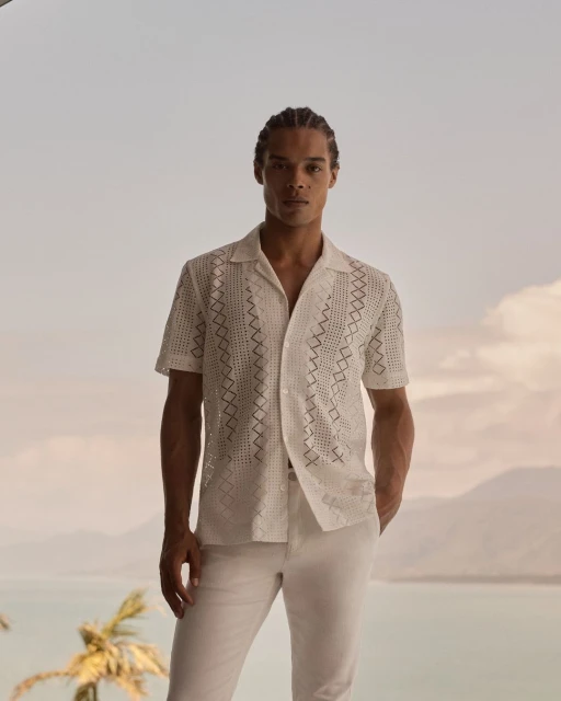 Man in white patterned shirt and pants stands outdoors with a calm expression, showcasing a summer fashion style and tropical backdrop.