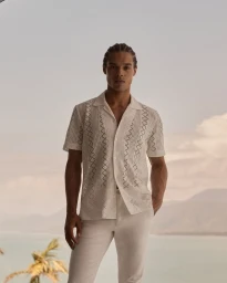 Man with braided hair wearing a white patterned short-sleeve shirt and matching trousers against an outdoor coastal background.