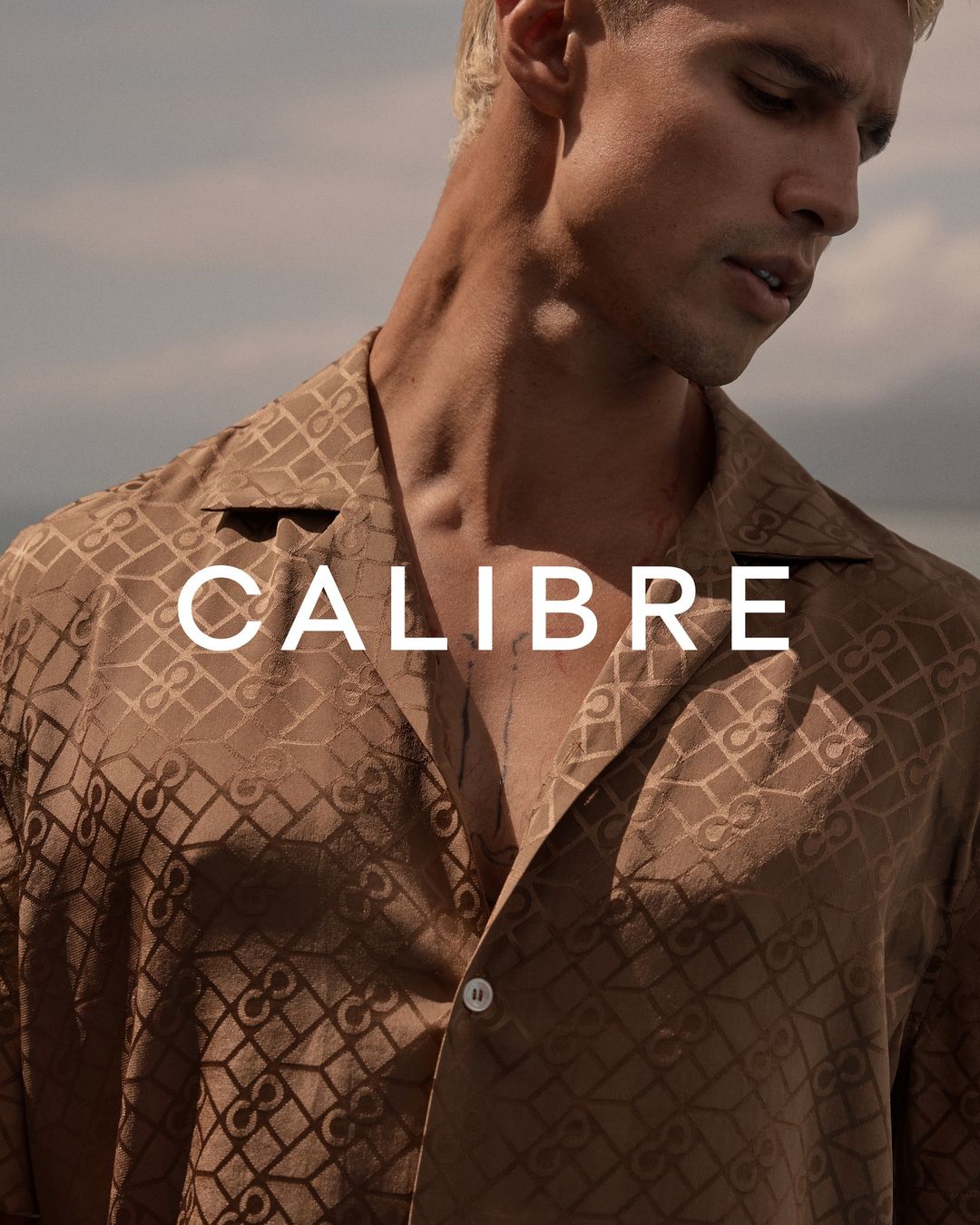 Portrait of a light-skinned male model with platinum blond hair wearing a patterned button-down shirt against a muted outdoor background.