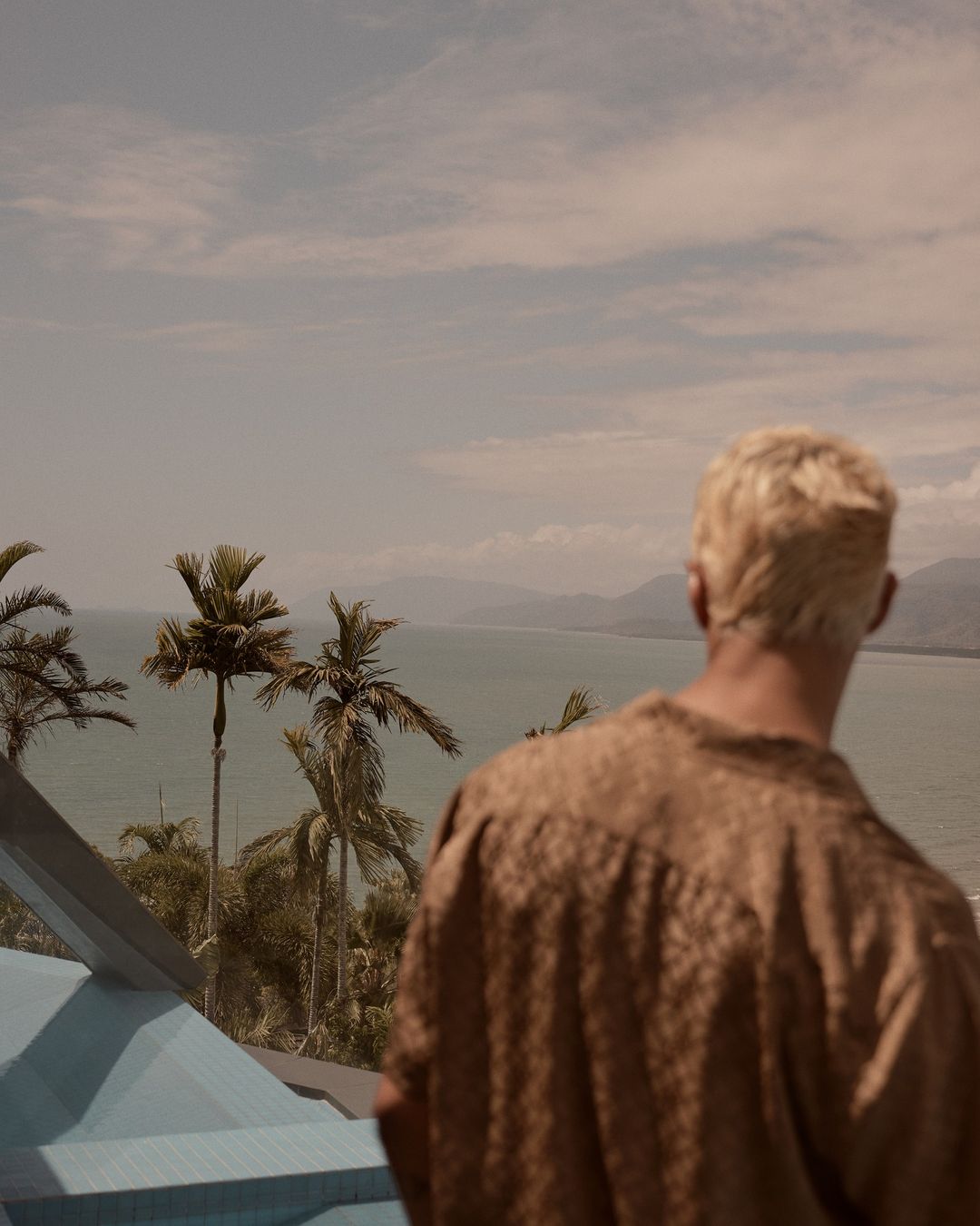 Blonde man gazes at distant mountains and ocean framed by palm trees; captured from behind in a serene, tropical outdoor setting.
