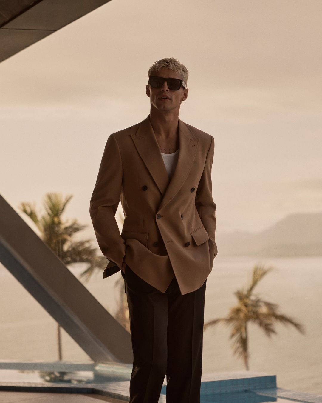 Adult male in a tan suit and sunglasses stands on a patio overlooking the ocean with palm trees in the background in a portrait shot.