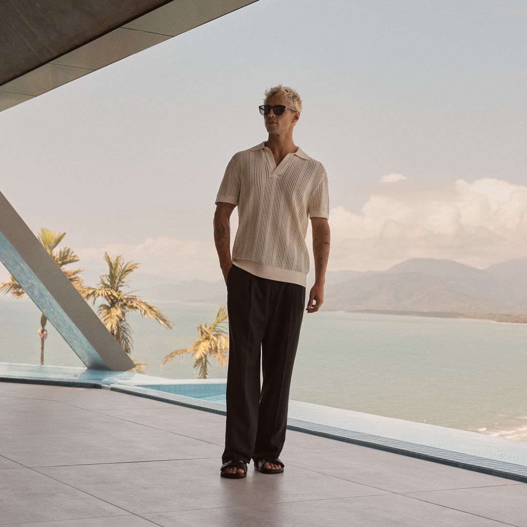 Male fashion model in polo shirt and slacks stands on a terrace overlooking an ocean and tropical mountains on a sunny day.