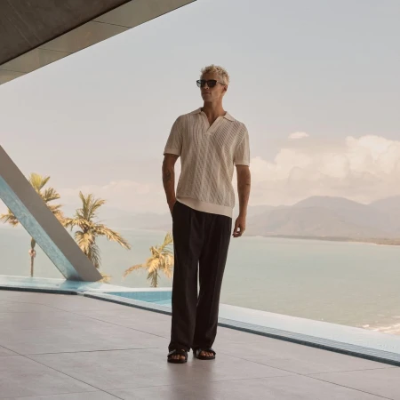 Male fashion model in polo shirt and slacks stands on a terrace overlooking an ocean and tropical mountains on a sunny day.