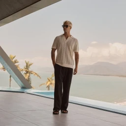 Man wearing sunglasses and a cream knit polo shirt standing on a modern outdoor terrace near a pool with palm trees.