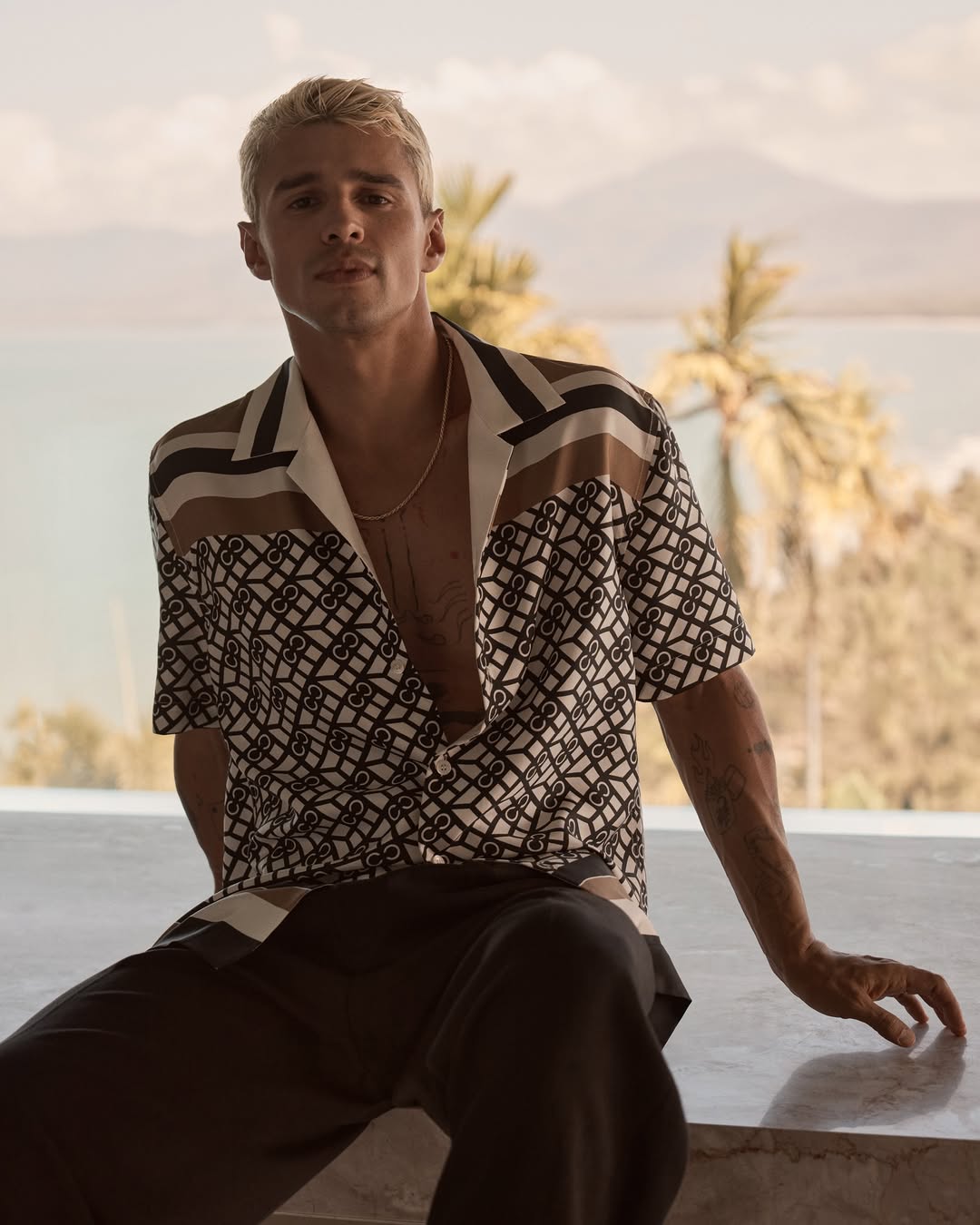 A blonde man in a patterned shirt sits on a ledge, with a scenic background featuring mountains, palm trees, and a body of water.