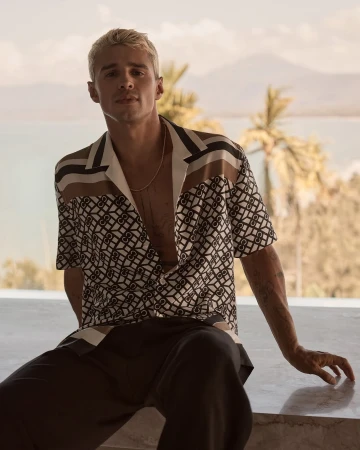 A blonde man in a patterned shirt sits on a ledge, with a scenic background featuring mountains, palm trees, and a body of water.