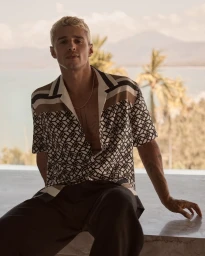 Blonde male model wearing a patterned short-sleeve resort shirt outdoors against a tropical coastal landscape background.