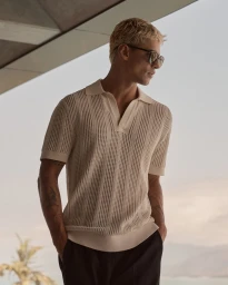 Blonde man wearing a cream knit polo shirt and sunglasses standing outdoors near a balcony overlooking a landscape