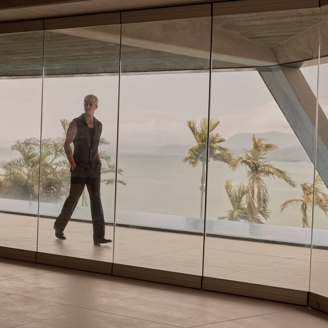 An adult male model in a dark outfit walks inside a modern home with a large glass window with a tropical landscape in the background.