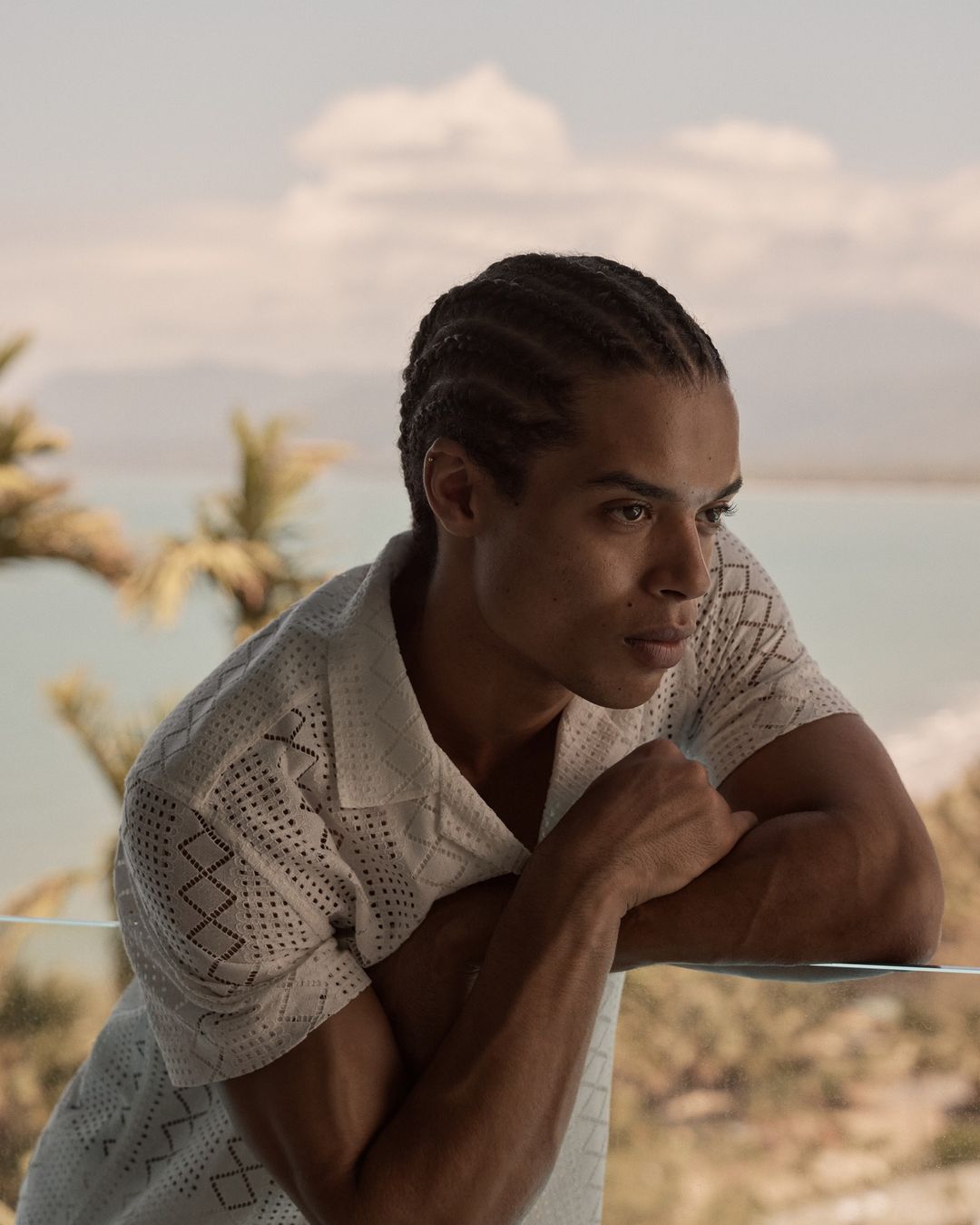 Portrait of a thoughtful-looking adult male with braided hair, wearing a short-sleeved patterned shirt, posing outdoors with a coastal view.