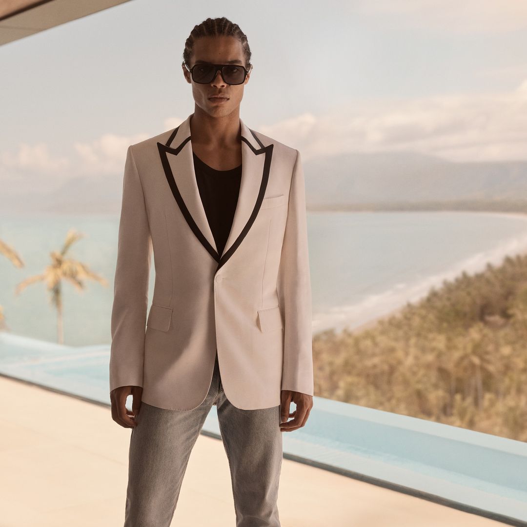 A male model stands outdoors near a pool, wearing a black T-shirt, a cream-colored suit jacket with black trim, and light gray jeans.
