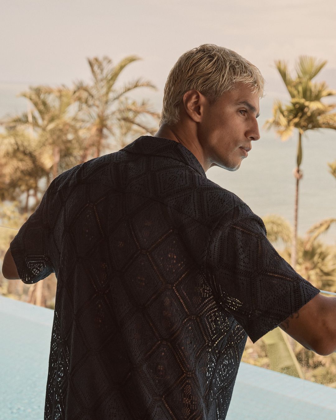 Close-up of an adult male in a patterned dark-blue shirt standing by a pool with a tropical ocean view in the background.