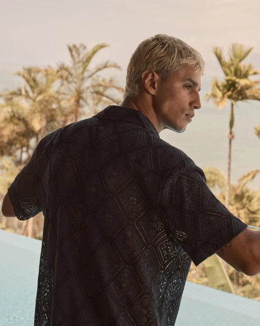 Close-up of an adult male in a patterned dark-blue shirt standing by a pool with a tropical ocean view in the background.