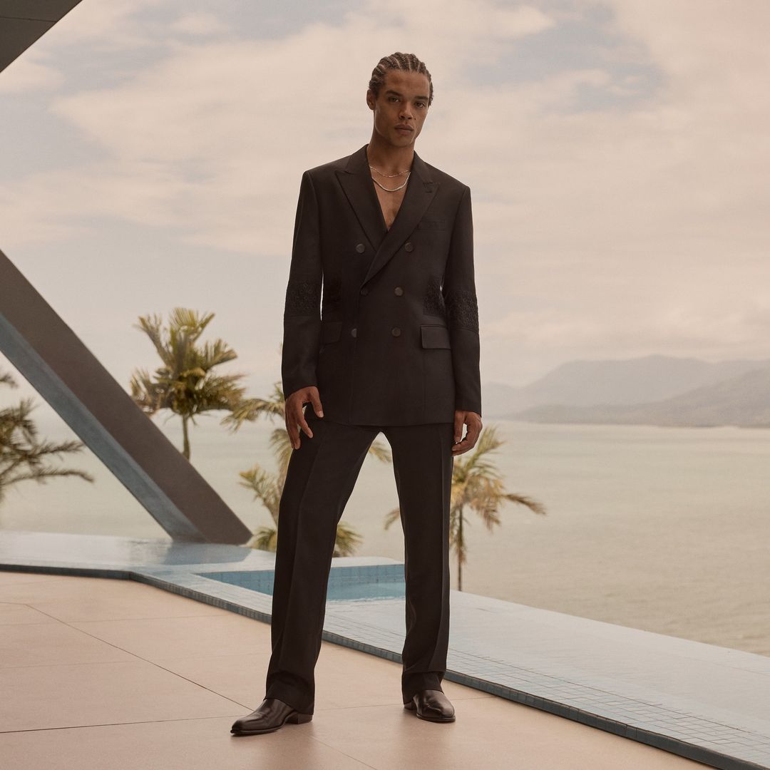 Man in a black suit stands outdoors against a backdrop of palm trees, a body of water, and cloudy sky, beside a modern pool.