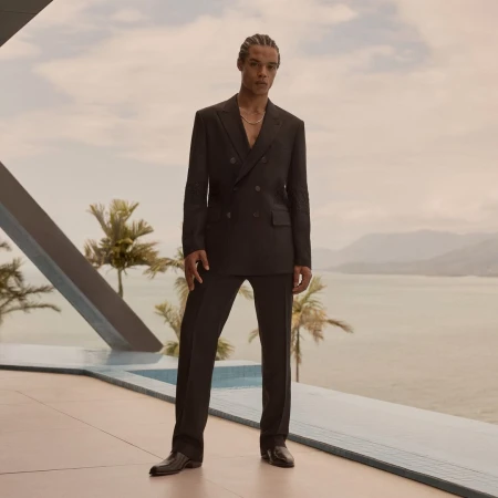 Man in a black suit stands outdoors against a backdrop of palm trees, a body of water, and cloudy sky, beside a modern pool.