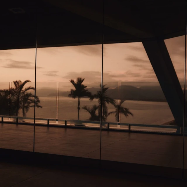 View of silhouetted palm trees and the ocean at sunset seen through large windows, creating a serene, tropical indoor-outdoor scene.