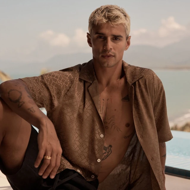A fair-skinned adult male with blonde hair poses in a brown patterned button-down shirt with a blurred background of water and land.