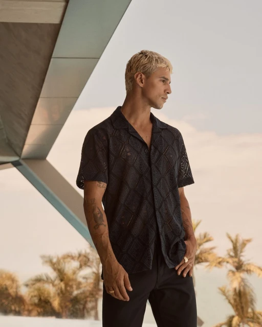 Man in black lace shirt and black pants poses outside, showcasing fashion and style with blurred trees and modern architecture backdrop.
