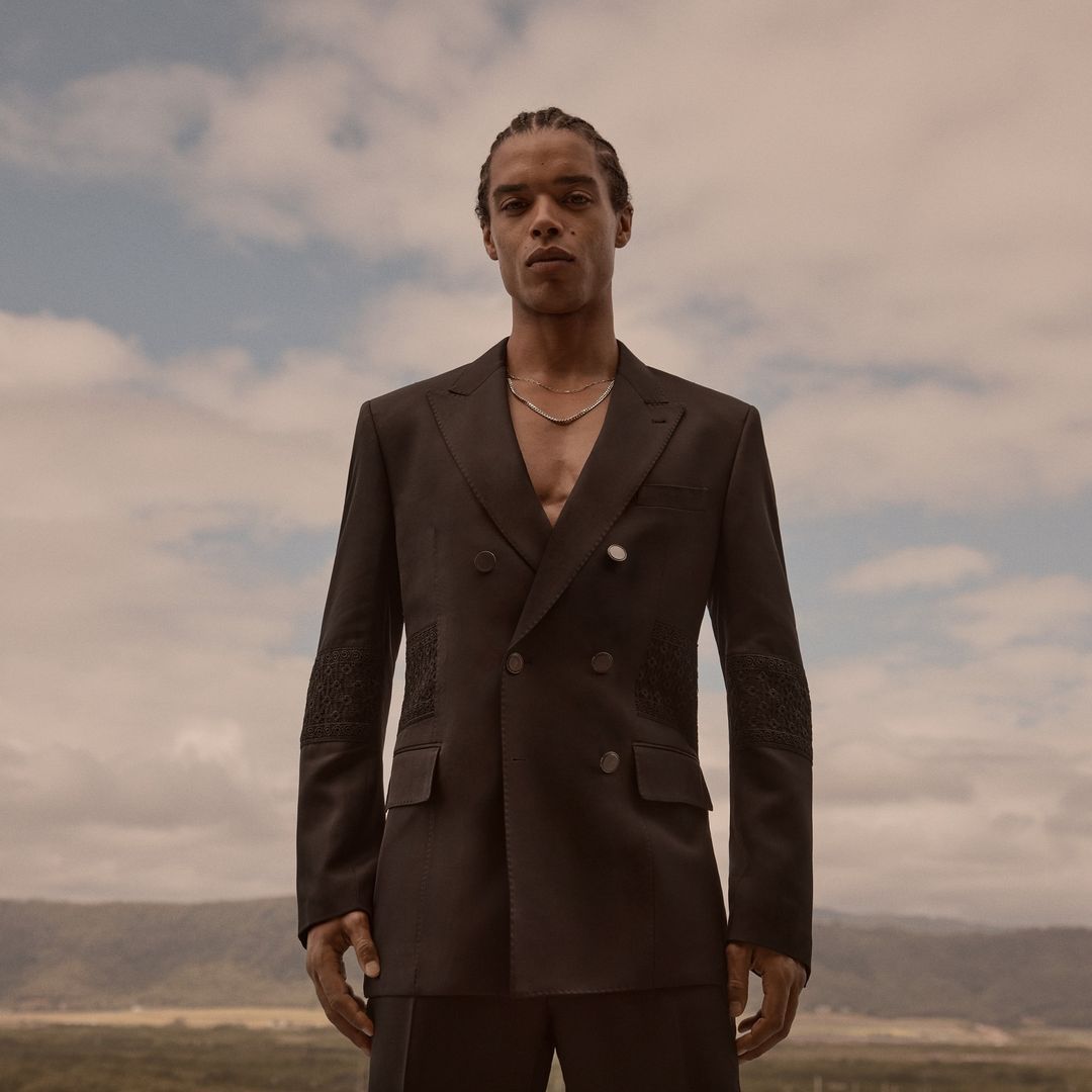 Man wearing a dark double-breasted suit jacket outdoors against a cloudy sky background for Calibre High Summer