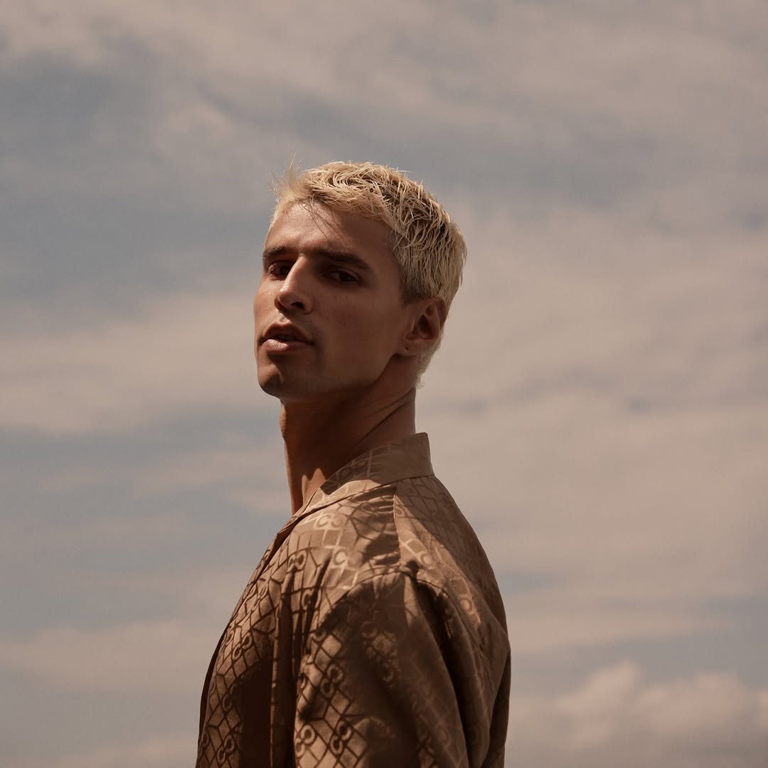 Blonde male model poses outdoors against cloudy sky wearing patterned button-down shirt in a low-angle portrait capturing fashion and style.