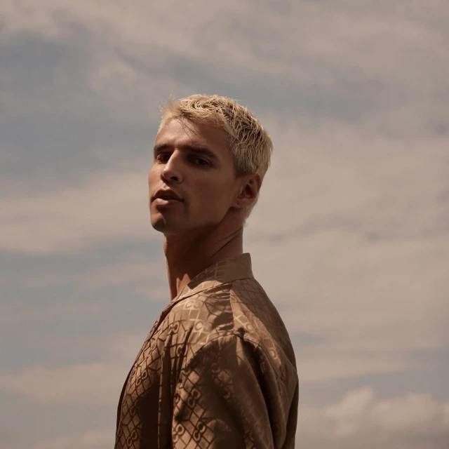 Blonde male model poses outdoors against cloudy sky wearing patterned button-down shirt in a low-angle portrait capturing fashion and style.